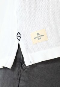 Polo blanco de manga corta con un diseño de punto texturizado, que presenta una etiqueta que dice "NO SOLO CUALQUIER MARCA" y un pequeño emblema cerca del dobladillo.