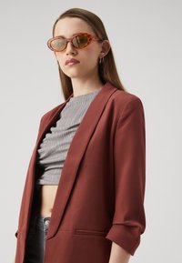 Lunettes de soleil avec des verres teintés ambre et des montures transparentes orange. Modèle portant un blazer couleur rouille sur un haut texturé gris.