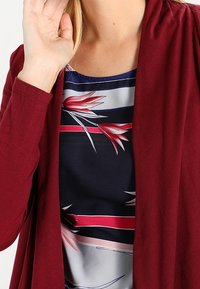 Burgundská cardigan s dlouhými rukávy přehozená přes modré tričko s květinovým a pruhovaným vzorem v červených, bílých a modrých tónech.