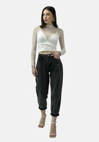 Hvid indpakningsbluse med gennemsigtige lange ærmer, højtaljede sorte jeans og klare ankelstrop hæle, sat mod en neutral baggrund.