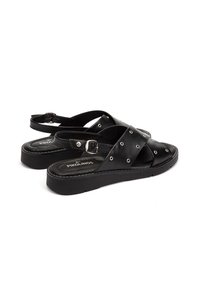 Sandalias de piel negra con tiras cruzadas, detalles de remaches plateados y correa de tobillo ajustable sobre una suela de plataforma baja. Nombre de la marca visible.