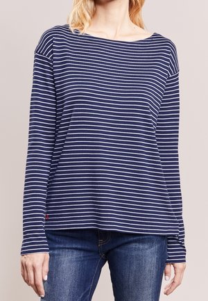 Vrouw draagt een marineblauw shirt met lange mouwen en witte horizontale strepen, en donkere blauwe spijkerbroek, staand tegen een effen achtergrond.