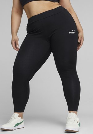 Czarne legginsy wykonane z elastycznego materiału, z wysokim stanem, z białym logo Puma na lewym udzie, w komplecie z sportowymi białymi butami.