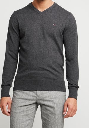 Mann mit dunkelgrauem V-Ausschnitt-Pullover und hellgrauen texturierten Hosen, steht mit entspannten Armen an den Seiten.