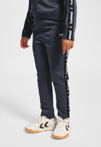 Granatowe spodnie dresowe wykonane z gładkiego materiału o zwężanym kroju, z białymi akcentami z logo po bokach. Połączone z białymi butami sportowymi.