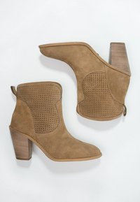 Stivaletti alla caviglia in suede beige con pannello laterale perforato, punta acuta e tacco in legno impilato. Presentano un'etichetta sul retro per un facile indossamento.