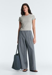 Femme portant un haut gris ajusté à manches courtes, pantalon large à carreaux, tongs noires, tenant un grand sac cabas foncé sur le côté.