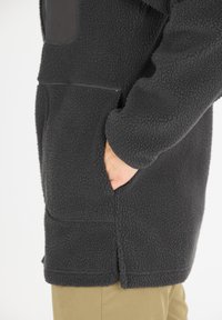 Fleece takki tummanharmaana, teksturoitua kangasta, jossa on suuri etutasku, sivuhalkio ja istuvat hihat. Minimalistinen muotoilu pehmeällä pinnalla.