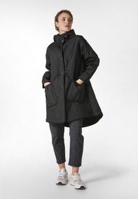 Sort quiltet frakke med hætte, med trykknapper og snøre i taljen, parret med grå tætsiddende bukser og hvide sneakers.