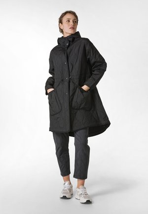 Zwarte gewatteerde jas met capuchon, voorzien van drukknoopsluitingen en een verstelbare taille, gecombineerd met grijze tapered broek en witte sneakers.