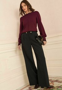 Burgundská halenka s dlouhým rukávem a výstřihem s vlnkovým lemováním, doplněná širokými černými kalhotami s vysokým pasem, na kterých jsou zlato-tonované knoflíky.