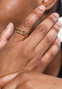 Ring i gulltone med sammenvevde bånd på en teksturert finger. Hånden hviler mot en lett solbrun hudoverflate.