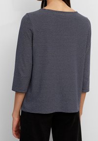 Eine Frau trägt ein navyblau-weiß gestreiftes Langarm-Shirt mit Dreiviertelärmel, von hinten gezeigt, kombiniert mit schwarzen Hosen.