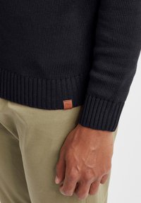 Maglione nero lavorato a maglia con polsini e orlo a coste, dotato di un piccolo tag in pelle marrone con logo. Indossato sopra pantaloni beige chiaro.