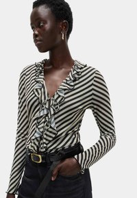 Donna che indossa una blusa a strisce diagonali bianche e nere con volant, cintura nera con fibbia dorata e pantaloni scuri, con una mano in tasca, che guarda di lato.