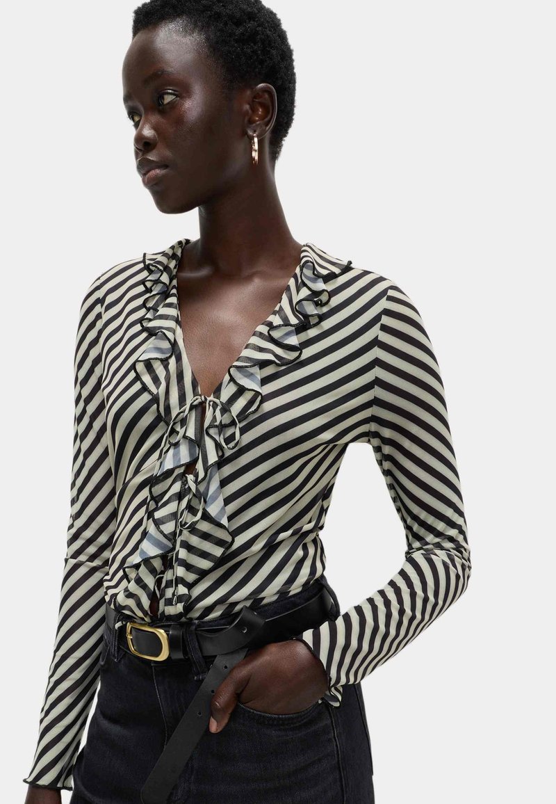 Donna che indossa una blusa a strisce diagonali bianche e nere con volant, cintura nera con fibbia dorata e pantaloni scuri, con una mano in tasca, che guarda di lato.