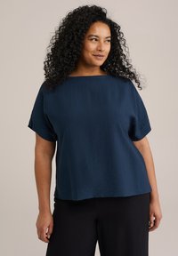 Top de manga corta azul marino con forma cuadrada y un escote amplio, fabricado en un tejido ligero con una sutil textura. Combinado con pantalones negros.