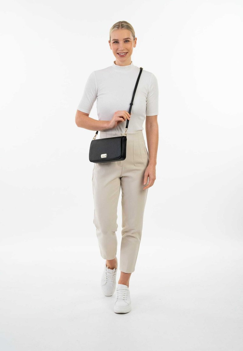 Zwarte leren crossbody tas met een rechthoekige vorm, gouden hardware en een afneembare schouderband. Model draagt een witte top en beige broek.