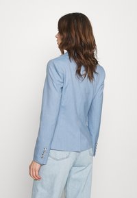 Ljusblå skräddarsydd blazer med en figursydd design, texturerad tyg och silverfärgad knappräkning på ärmarna. Bärs över ljus denim.