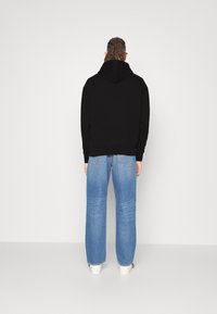 Schwarzer Hoodie mit Kängurutasche, kombiniert mit hellblauen, geraden Jeans. Weiße Sneaker sichtbar, vor einem sauberen, minimalistischen Hintergrund.