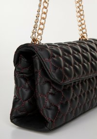 Bolso de mano de piel negra con textura acolchada de patrón de corazones, con costuras rojas y una correa de cadena dorada. Forma rectangular compacta.