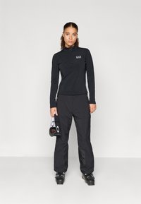 Čierny zipsový top s dlhým rukávom a logom EA7, v kombinácii s čiernymi nepremokavými nohavicami. Model drží čiernu tašku na lyžiarske topánky s bielymi akcentmi.