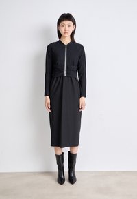 Schwarz Kleid mit einem taillierten, mit Reißverschluss versehenen Oberteil und plissiertem Rock. Lange Ärmel und strukturierter Stoff. Getragen mit knielangen schwarzen Stiefeln.