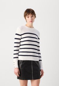Hvit strikkegenser med marineblå horisontale striper, ribbestrikket halslinning og mansjetter, med dekorative knappdetaljer på ermet. Kombinert med et sort skinnskjørt.