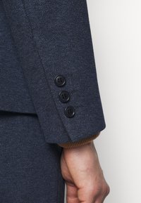 Giacca di abito blu navy con polsino su misura e tre bottoni neri, tessuto testurizzato e un accento lavorato a maglia marrone visibile sotto la manica.