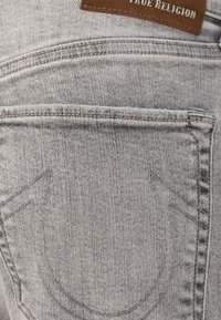 Jasnoszare dżinsy z wyraźną fakturą, z kontrastującą brązową skórzaną łatą z napisem "TRUE RELIGION" oraz szczegółowym szyciem.