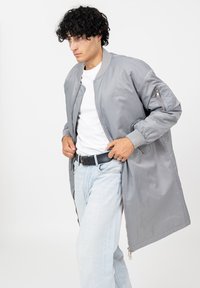 Man met krullend zwart haar draagt een lange grijze bomberjack, een wit T-shirt, lichtblauwe spijkerbroek en een zwarte riem, terwijl hij de jas bij de taille rechtzet.