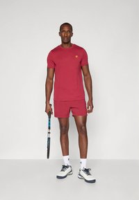 Mees punases spordiriietuses, tennisereket käes, valgete sokkide ja valgete tennistega, seisab tühjal taustal.