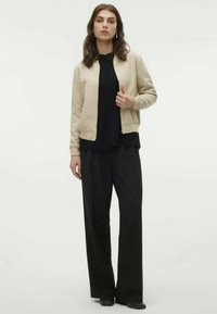 Light beige bomber jacket with ribbed cuffs, worn over a black layered top and wide black trousers, featuring a smooth texture and casual fit.