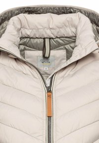 Beige Steppjacke mit Reißverschluss, mit gefüttertem Kragen und Zickzacksteppung. Mit einem Etikett für "Camel Active" und Größe 36.