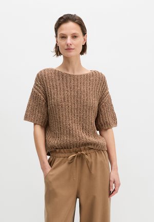 Woman with short brown hair wearing a loose brown knit short-sleeve top and tan drawstring pants with hands in pockets.