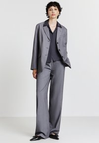 Giovane in piedi con un completo grigio a righe sottili, camicia nera e scarpe nere a punta, su sfondo bianco semplice.