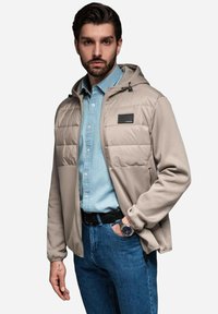 Ljusbeige jacka med huva, med vadderade frampaneler och en slät textur. Bärs över en denimskjorta med blå jeans.