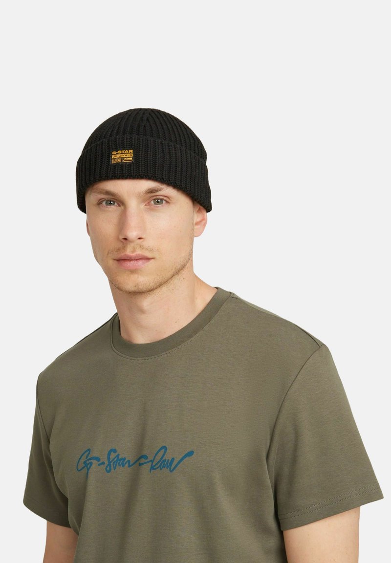 Berretto nero a costine con un piccolo etichetta gialla che mostra il nome del marchio. Indossato con una maglietta verde oliva che presenta del testo blu.