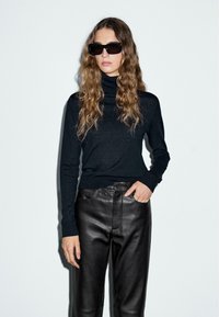 Donna che indossa un dolcevita nero, pantaloni di pelle nera e grandi occhiali da sole neri, in piedi contro uno sfondo chiaro e semplice.