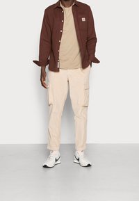 Brun långärmad skjorta över en beige t-shirt, som matchas med beige cargo-byxor och vita sneakers med svarta detaljer.