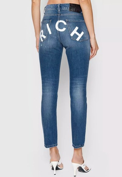 Vista posteriore di jeans skinny blu con dettagli sbiaditi, lettere bianche in blocco "RICH" sulle tasche posteriori, indossati con sandali bianchi con tacco alto.