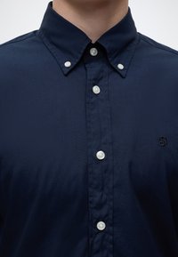 Gros plan d'une personne portant une chemise bleu marine boutonnée avec un col boutonné et un petit logo brodé sur la poitrine.