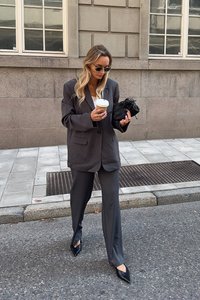 Siv kostim z valjnim blejzerjem in širokimi hlačami. Nosi se z ostrimi črnimi čevlji, drži skodelico kave in majhno črno torbico.