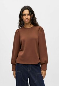 Braunes Longsleeve-Oberteil mit gerafften Puffärmel, Rundhalsausschnitt und glatter Textur, kombiniert mit dunkelblauen weit geschnittenen Jeanshosen.