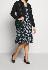 Czarna dżinsowa kurtka na czarnej sukience w kwiaty z niebieskimi i białymi wzorami, w połączeniu z czarnymi szpilkami i zieloną torebką.