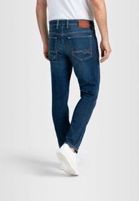 Dunkelblaue Denim-Jeans mit schmaler Passform. Mit Gesäßtaschen, einem braunen Lederpatch am Bund und weißen Sneakers.