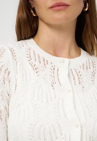 Γυναίκα που φοράει λευκό πλεκτό ζακέτα με κουμπιά από πέρλες και χρυσούς κρίκους στα αυτιά, εμφανίζοντας μόνο τον λαιμό και το κάτω μέρος του προσώπου.