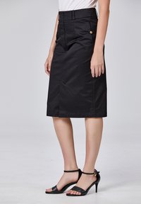 Jupe noire longueur genou faite d'un tissu lisse, avec une fente avant, des poches latérales et des accents en métal doré, associée à des sandales à talons noires.