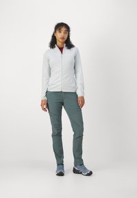 Lichtgrijze ritsjack met een aansluitend ontwerp, gecombineerd met donkergroene tapered broek en blauwe robuuste schoenen. Minimale branding zichtbaar.