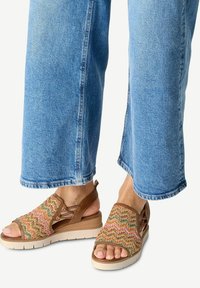 Sandali intrecciati con motivo a zigzag multicolore, accentuati da pelle color cuoio, con retro chiuso e suola piatta beige. Jeans parzialmente visibili.